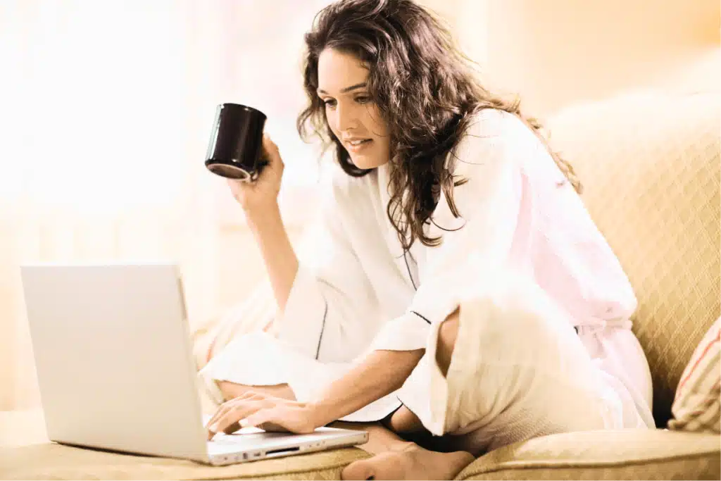 Woman in robe sitting on couch with coffee and laptop – relaxed morning work-from-home routine